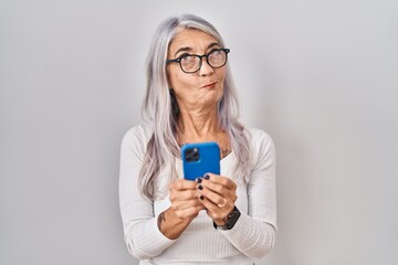 Middle age woman with grey hair using smartphone typing message smiling looking to the side and staring away thinking.