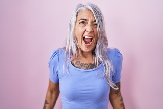 Middle age woman with tattoos standing over pink background angry and mad screaming frustrated and furious, shouting with anger. rage and aggressive concept.