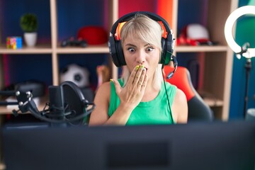 Young caucasian woman playing video games wearing headphones covering mouth with hand, shocked and afraid for mistake. surprised expression