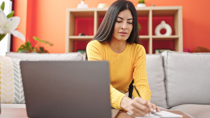 Young beautiful hispanic woman using laptop writing on notebook at home