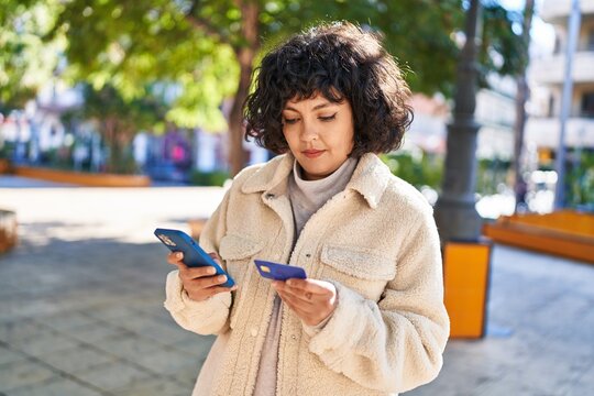 Young beautiful hispanic woman using smartphone and credit card at park