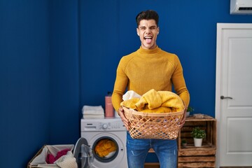 Young hispanic man holding laundry basket sticking tongue out happy with funny expression.