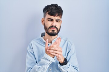 Young hispanic man with beard standing over blue background suffering pain on hands and fingers, arthritis inflammation