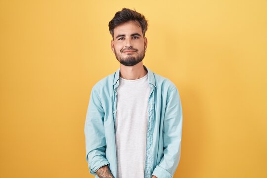 Young hispanic man with tattoos standing over yellow background with hands together and crossed fingers smiling relaxed and cheerful. success and optimistic