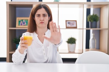 Brunette woman drinking glass of orange juice doing stop sing with palm of the hand. warning expression with negative and serious gesture on the face.