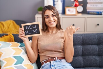 Young hispanic woman holding blackboard with new home text pointing thumb up to the side smiling happy with open mouth