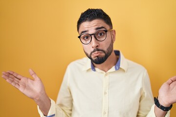 Hispanic young man wearing business clothes and glasses clueless and confused expression with arms and hands raised. doubt concept.