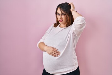 Pregnant woman standing over pink background confuse and wondering about question. uncertain with doubt, thinking with hand on head. pensive concept.