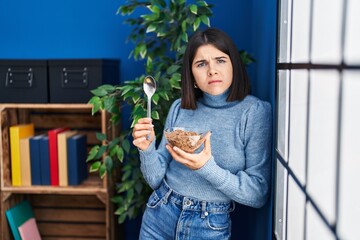 Young hispanic woman eating healthy whole grain cereals with spoon clueless and confused...