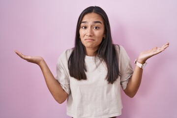 Young hispanic woman standing over pink background clueless and confused expression with arms and hands raised. doubt concept.