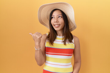 Middle age chinese woman wearing summer hat over yellow background smiling with happy face looking and pointing to the side with thumb up.