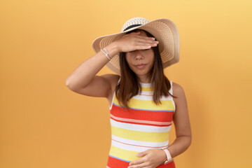 Middle age chinese woman wearing summer hat over yellow background covering eyes with hand, looking serious and sad. sightless, hiding and rejection concept