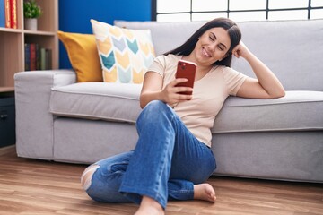 Fototapeta premium Young beautiful hispanic woman using smartphone sitting on floor at home