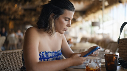 Young hispanic woman using smartphone sitting on the table at the restaurant