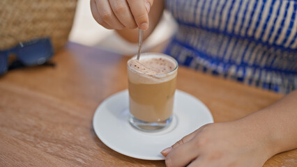 Young hispanic woman stirring coffee sitting on the table at sunny restaurant terrace