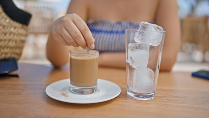 Young hispanic woman stirring coffee sitting on the table at sunny restaurant terrace