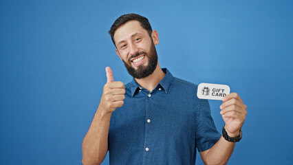 Young hispanic man smiling confident holding gift card doing thumb up gesture over isolated blue...