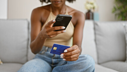 African american woman make photo to credit card by smartphone sitting on sofa at home