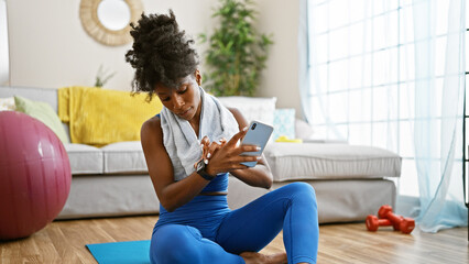 African american woman wearing sportswear looking watch and smartphone at home