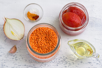 Ingredients for vegan tomato lentil sauce, soup or for lentil pancakes: red lentils, onions, garlic, canned tomatoes, spices and vegetable oil on a light gray textured background