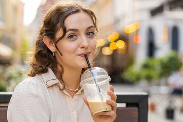 Happy young woman enjoying morning cold coffee drink with ice and smiling outdoors. Relaxing, taking a break. Girl sits on bench on city sunny street, drinking coffee to go. Town lifestyles outside