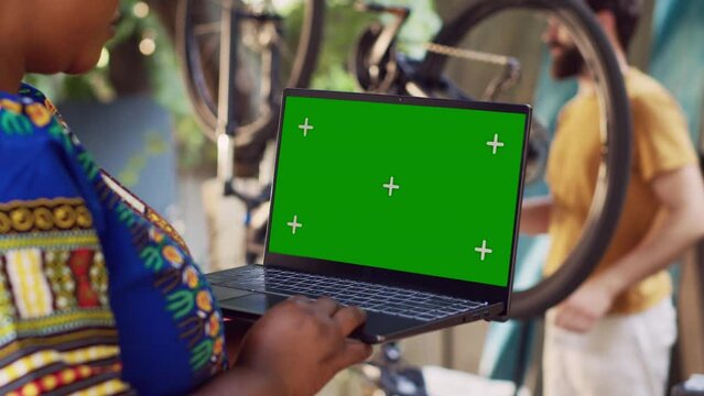 Detailed view of african american lady grasps minicomputer with greenscreen display as man in background removes broken wheel. Black woman holds laptop with blank mockup screen for bike maintenance.