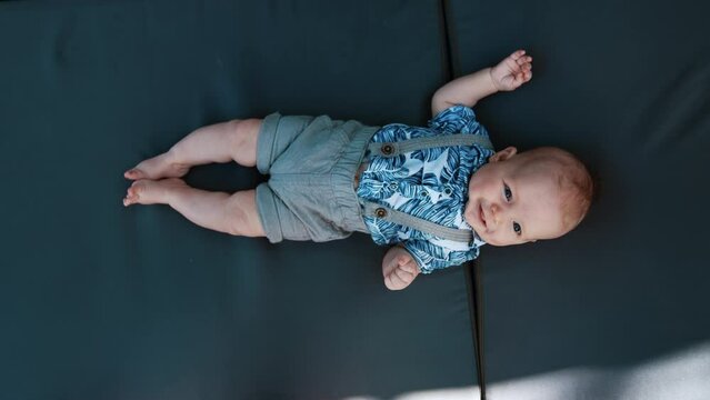 Adorable Caucasian baby boy wearing a blue shirt and shorts with suspenders. Smiling infant moving cheerfully his feet and hands. Vertical screen close up.