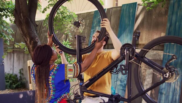 Dedicated boyfriend and girlfriend doing annual maintenance on bike tires for safe cycling in home yard. Youthful multiracial pair successfully mounting wheel on modern bicycle frame.