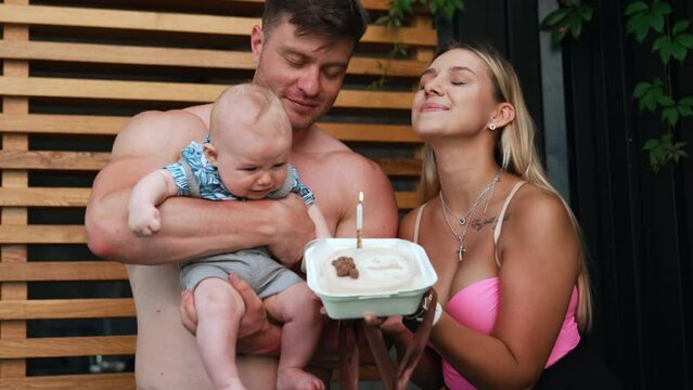 Family of three celebrating kid's month anniversary with a cake. Happy parents are blowing a candle and kid is starting to cry.