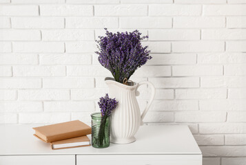 Jug with beautiful lavender flowers and books on chest of drawers near light brick wall in room, closeup