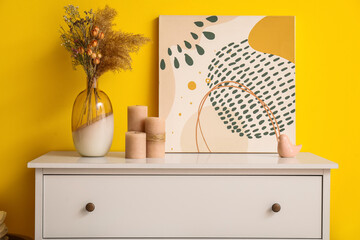 Vase with dried plant and painting on white chest of drawers near yellow wall