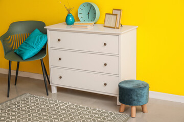 White chest of drawers and stylish chair with cushion near yellow wall