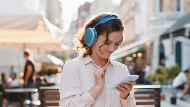 Happy relaxed overjoyed pretty young woman in wireless headphones choosing, listening favorite energetic disco rock n roll music dancing outdoors. Girl walking in urban sunshine city street background