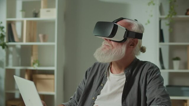 A Modern Old Man With Gray Beard in VR Glasses Examines Everything Around. An Elderly Man in VR Glasses Sitting at a Table with a Laptop. VR for the Socialization of the Elderly.