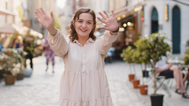 Hello. Young pretty woman smiling friendly at camera, waving hands gesturing hi, greeting or goodbye, welcoming with hospitable expression outdoors. Happy lovely girl walking in urban city town street
