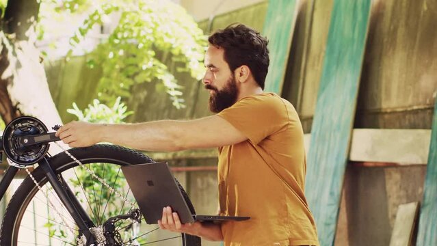 Male caucasian cyclist utilizing laptop to reasearch for online repair guides while repairing broken bicycle pedal. Healthy young man upkeeping bike by surfing the internet on minicomputer.