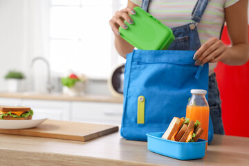 Young woman putting school lunchbox into backpack in kitchen
