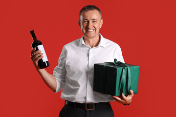 Mature man with bottle of wine and gift on red background