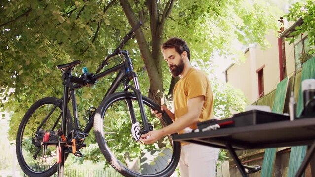 Sports-loving male loosening and removing damaged wheel from bike front fork with expert multitool outside. Committed man examining bicycle handlebar stem and headset in yard. Dolly zoom-in.