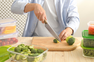 Man cutting fresh broccoli with knife near containers at wooden table, closeup. Food storage