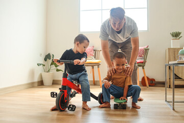 Happy young Asian father enjoying playing with sons at home. Children play with tricycle and skateboard. 