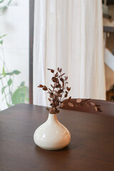 Dried leaf in ceramic vase on wooden table with white curtain background. Modern stylish home decor interior. Scandinavian neutral trendy colors.