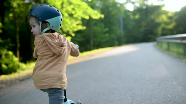 Funny toddler boy riding a baby scooter outdoors on summer day. Summer outdoor activities for kids. Slow motion footage.