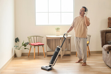 Happy young man wearing wireless headphones cleaning home with vacuum cleaner.