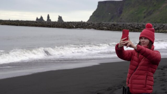 The girl takes pictures of the ocean on the phone. The girl is standing on a black beach. Iceland.