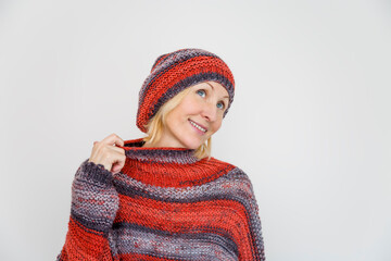 A woman dressed in a knitted terracotta sweater and a beanie pulls the collar of the sweater aside with her finger.