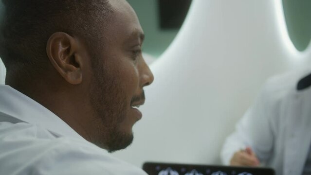 Close up of African American doctor laughing, talking with colleagues during lunch in clinic cafe. Digital tablet with MRI brain scan image. Medical staff in hospital or medical center cafeteria.