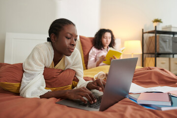 Fototapeta premium Black teenage girl working on laptop when friend reading book in background