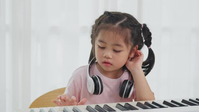 Asian cute girl smile and wearing white headphone playing learning online piano music in the living room at home. The idea of activities for the child at home during quarantine. Music learning study.