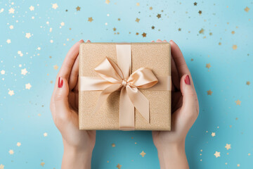 The photo shoot of a woman hands holding a wrapped gift box by paper and ribbon isolated on light blue background with metallic star glitter confetti or bokeh light. Generative AI.